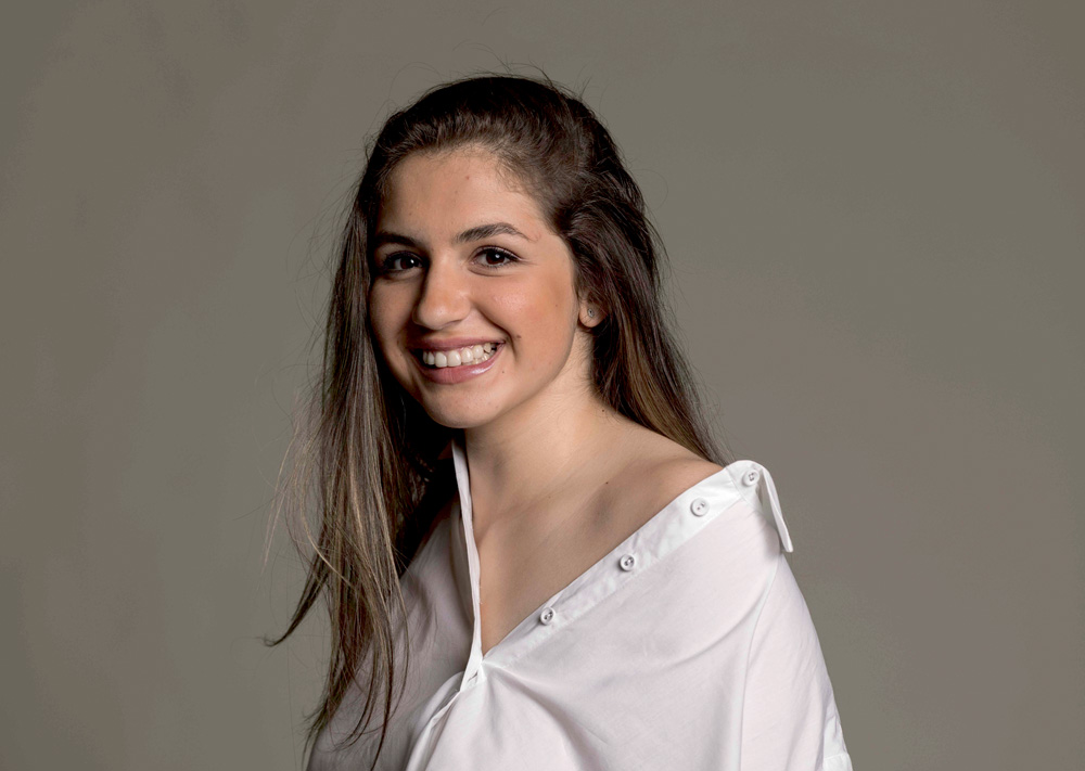 Isa Dervalli sorri para a foto e veste blusa branca. Tem cabelos longos escuros.