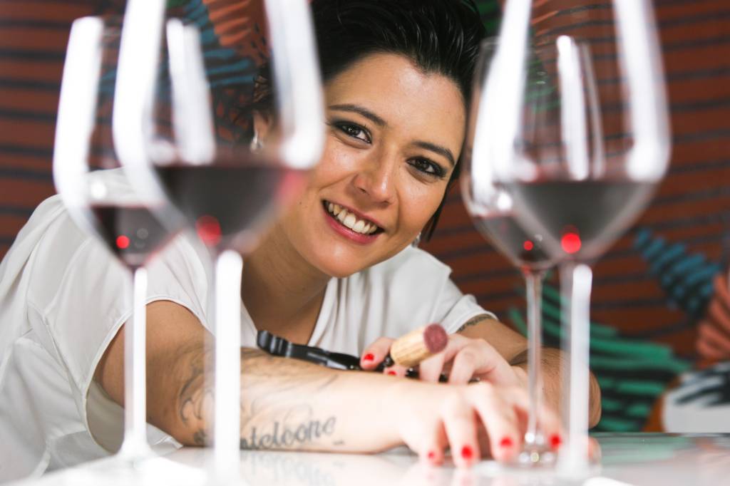 Tão Longe, Tão Perto Gabriela Monteleone posa sorridente entre taças de vinhos