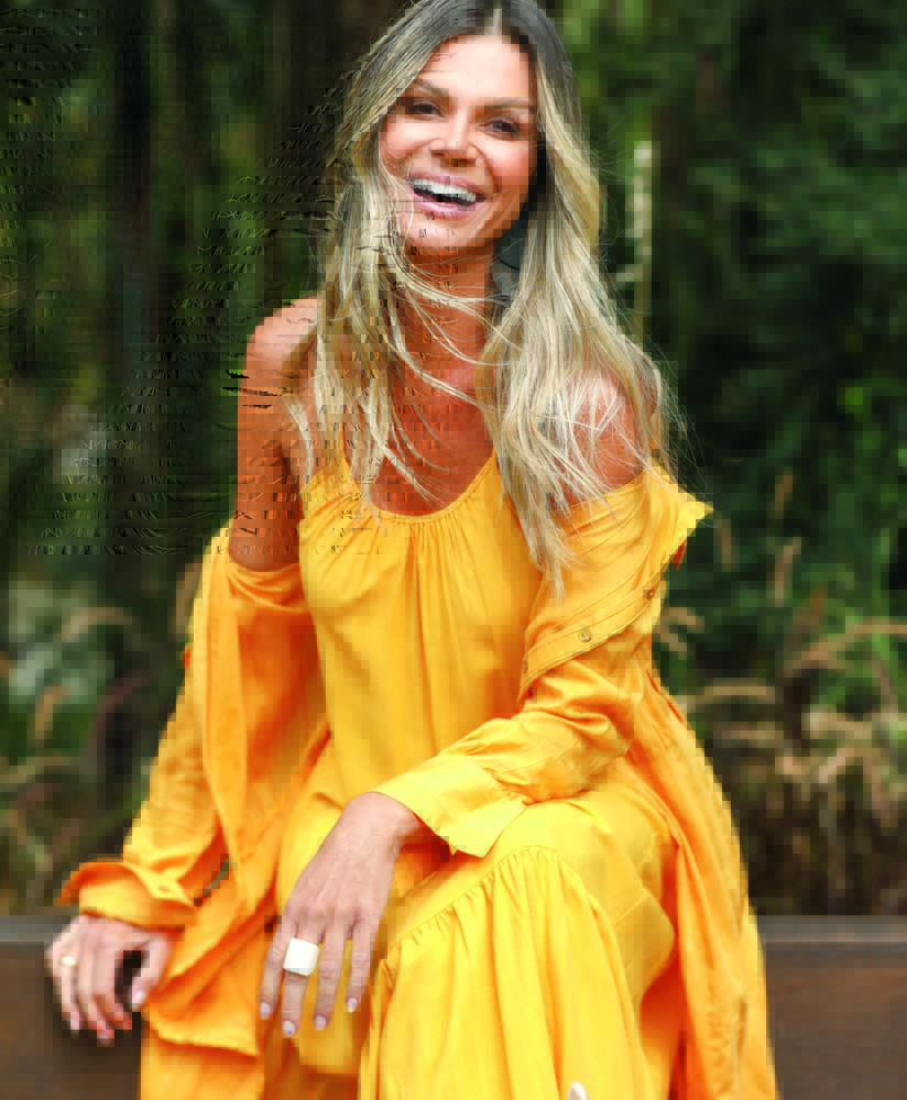 2021-06-02 14_38_33.318-0300.jpg Denize é uma mulher branca e magra, de cabelos lisos e loiros. Ela está sentada, usa um longo vestido laranja e sorri para a foto