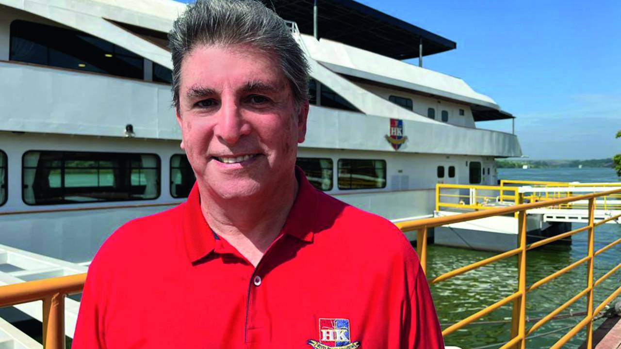 Imagem de Carlos Nascimento usando uma camiseta vermelha. Um barco está parado ao fundo da foto, na água.