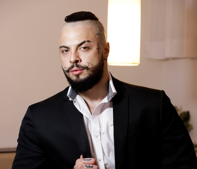 Gabriel Spec posa com blazer preto. Ele tem tatuagens no rosto, um moicano e bigode curvado.