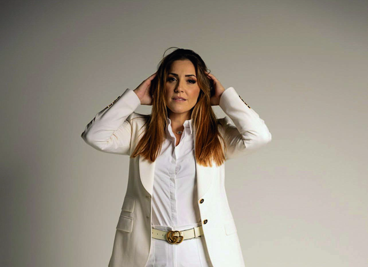 Thais &eacute; uma mulher branca, de cabelos e olhos castanhos. Ela usa uma camiseta, uma calca e um blazer pretos. Da cintura para cima, ela &eacute; fotografada em um fundo branco, com as m&atilde;os na cabe&ccedil;a