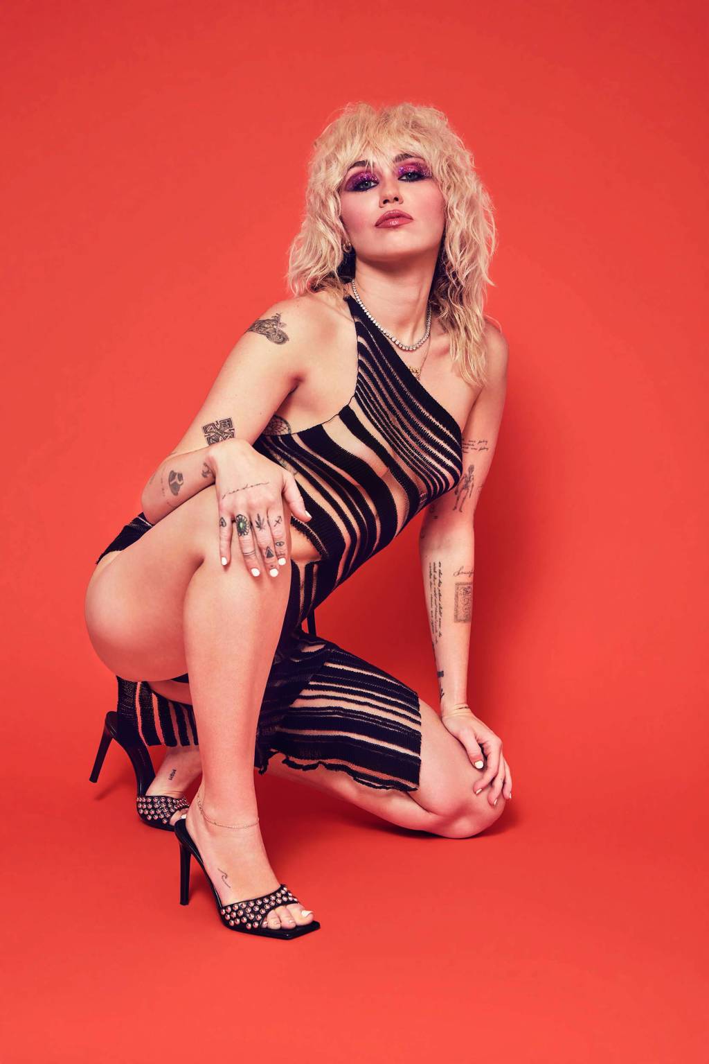 Miley Cyrus_divulgação.jpg Imagem mostra mulher de vestido agachada sobre fundo laranja.