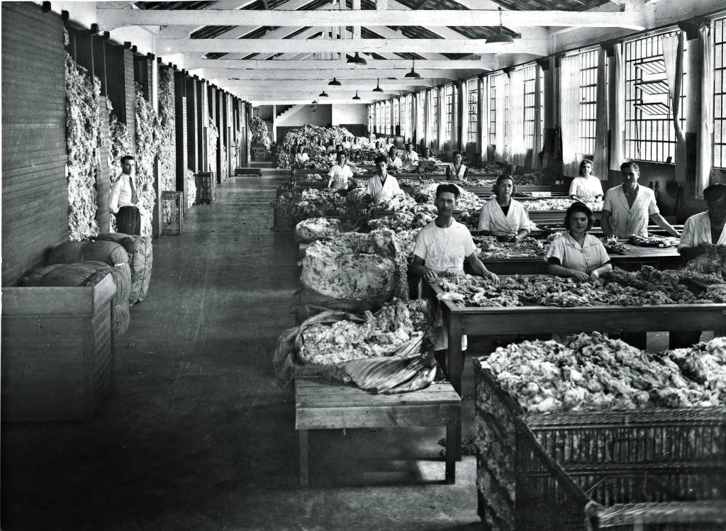 Sala de classificação de lã bruta da Fiação Santista Imagem mostra pessoas trabalhando em galpão com sacos de tecidos.