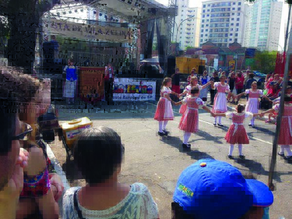 Festa da Vila Zelina Imagem mostra ciranda de crianças em área aberta. É possível ver um palco ao fundo, com um cartaz escrito