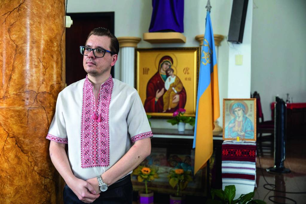 Vila Zelina – estefano Wonsik Imagem mostra padre apoiado em pilar de mármore. Ao fundo, uma imagem de Jesus.