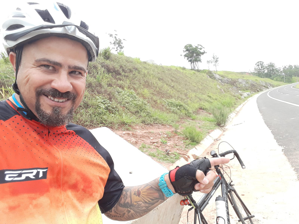 WhatsApp Image 2022-03-11 at 15.47.28 Luis é um homem negro, de barba. Ele usa um capacete e uma roupa adequada para ciclistas enquanto segura sua bicicleta em um trecho do rodoanel