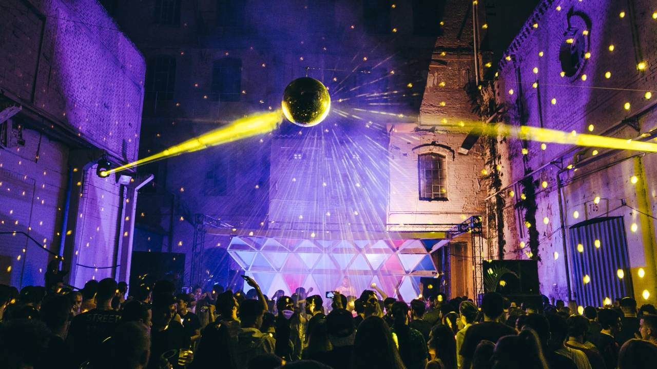 Imagem mostra pessoas em espaço aberto com luzes coloridas e um globo espelhado ao centro.