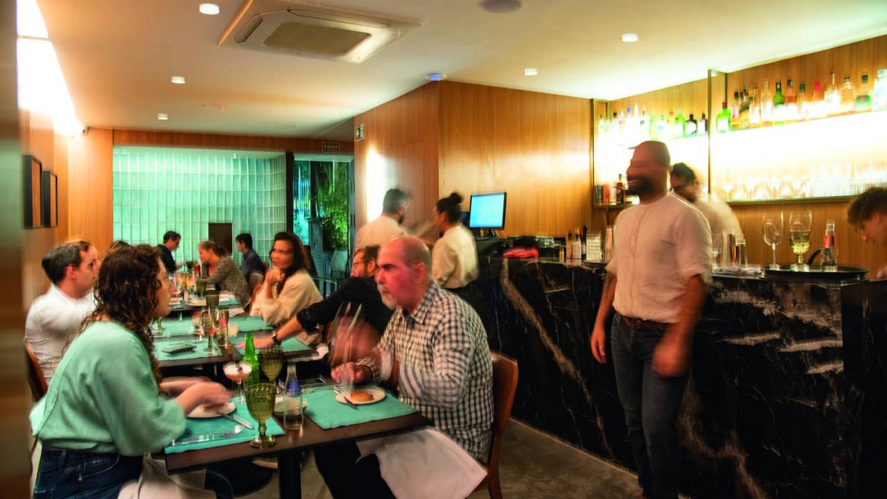 Ambiente de restaurante iluminado por brancas. Mesas são dispostas lado a lado em frente à parede e, ao fundo, há um balcão de mármore escuro onde encontra-se o bar, decorado por garrafas expostas em prateleiras