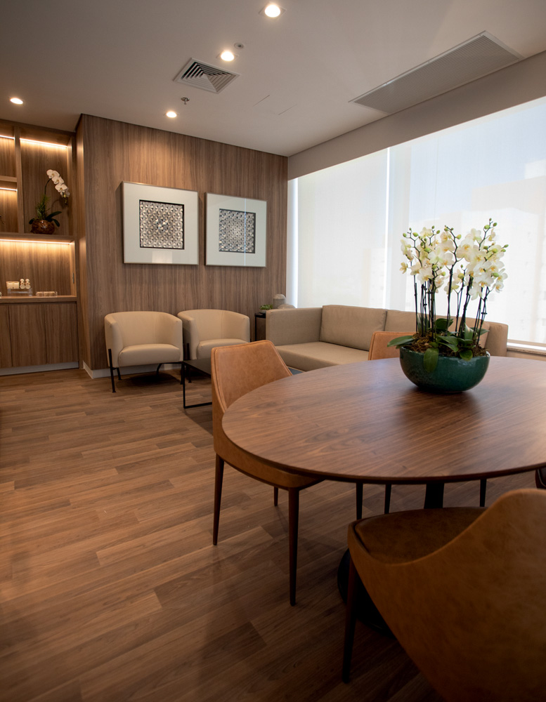 Quarto da maternidade em tons de marrom, com grande mesa de madeira redonda, poltronas, quadros e prateleiras iluminadas