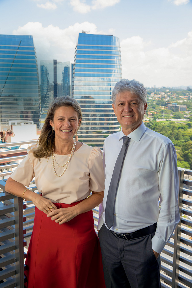 MATERNIDADE SÃO LUIz STAR Maria Augusta e Jair, que cuidam do novo empreendimento, aparecem na sacada do prédio. Ambos são brancos e de meia idade. Ela usa uma blusa salmão, uma saia vermelha e um colar de pérolas, tem cabelos lisos e castanhos. Ele é grisalho e usa uma camisa azul com gravata azul
