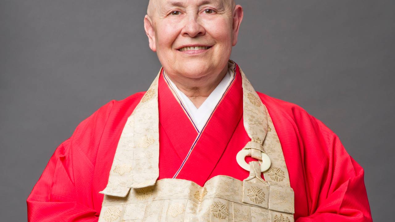 Monja Coen Roshi sorri para a foto com braços cruzados por baixo de roupa vermelha e peça em tom bege.