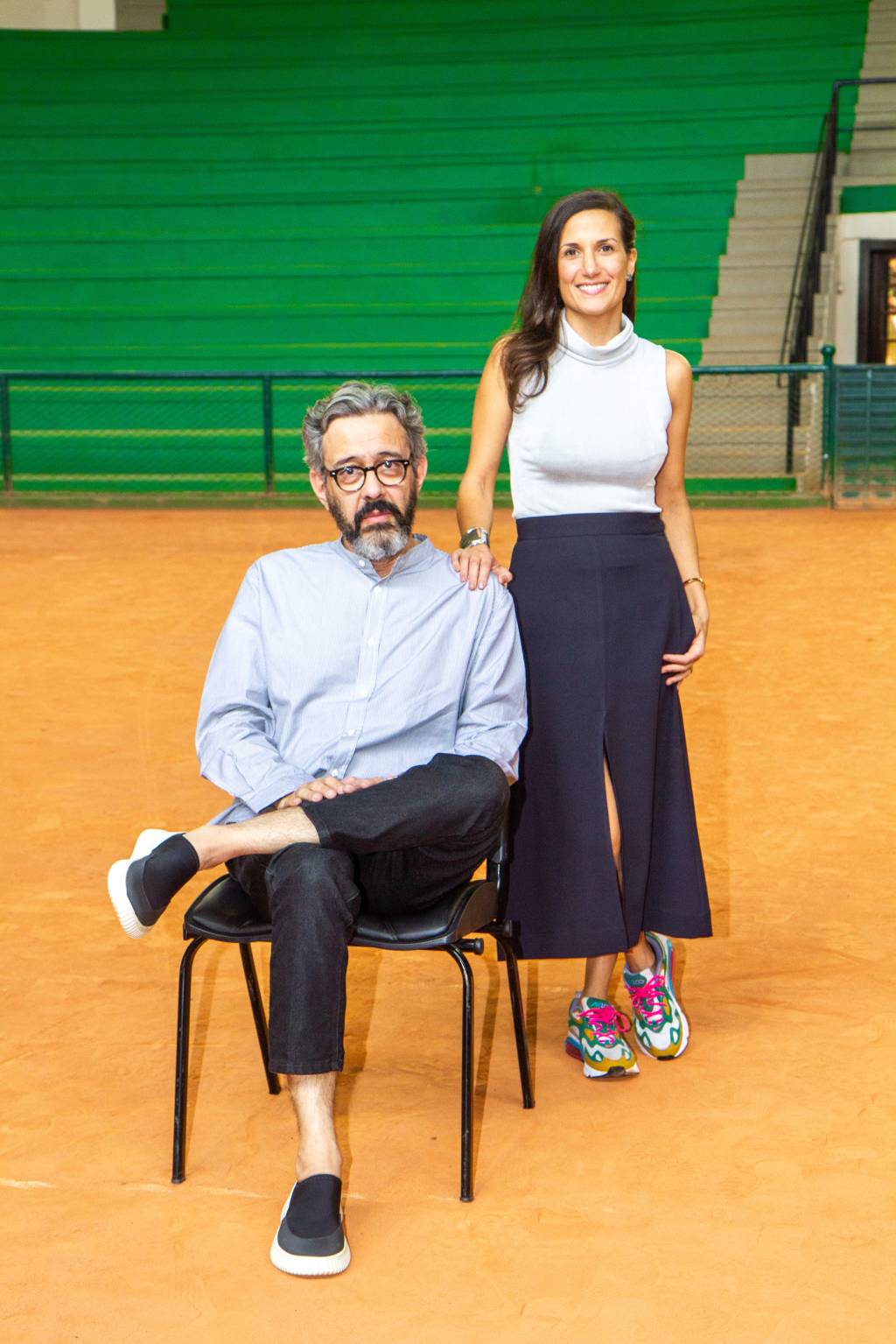 Waldick Jatobá e Camilla Barella. Crédito_ Silvana Garzaro(1) Waldick Jatobá e Camilla Barella, das feiras Made e ArPa, posam em um grande estádio com chão cor de areia. Ele está sentado de pernas cruzadas e ela de pé ao lado, com saia preta e blusa sem mangas branca.