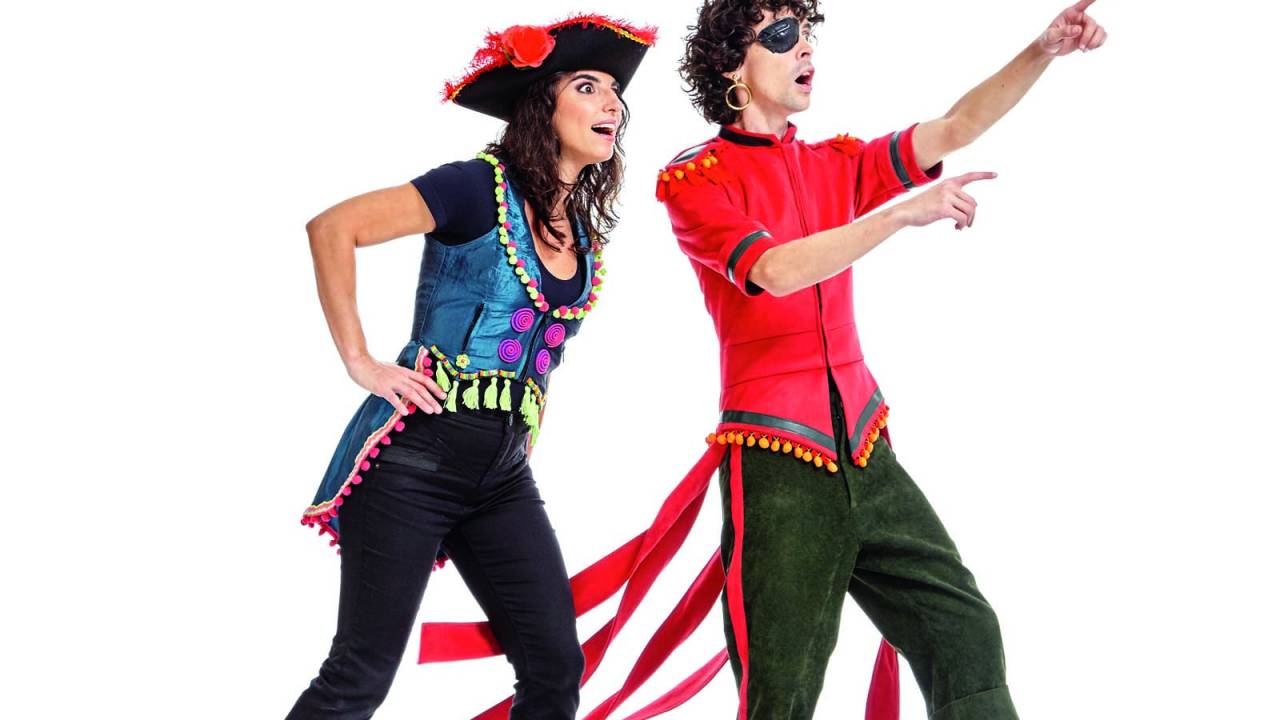 Julia e Daniel posam em um fundo branco, em cima de um tablado azul. Ambos usam fantasias divertidas de pirata e olham para um ponto cego na frente, com cara de impressionados