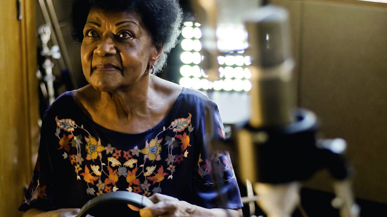 Imagem mostra mulher idosa segurando um fone. Um microfone é visto, desfocado, em primeiro plano.