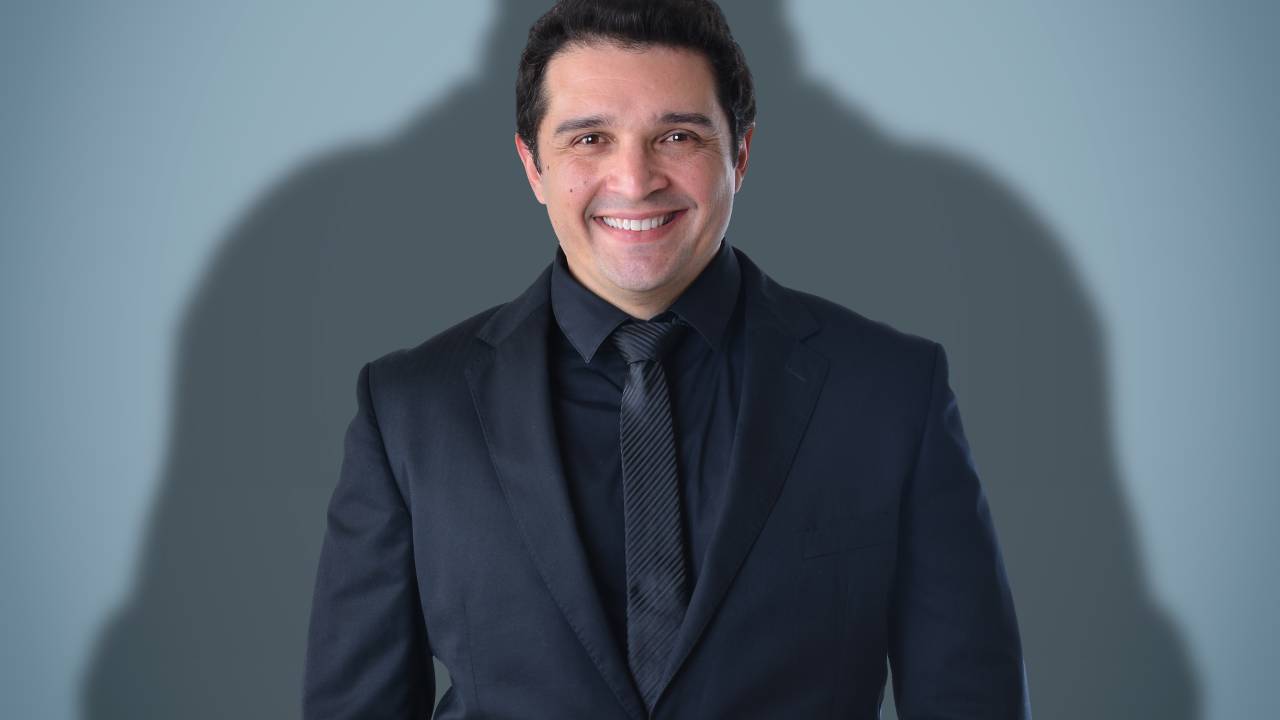 Homem branco posa de terno preto com silhueta de Batman logo atrás. Ele está sorrindo e tem cabelos pretos.