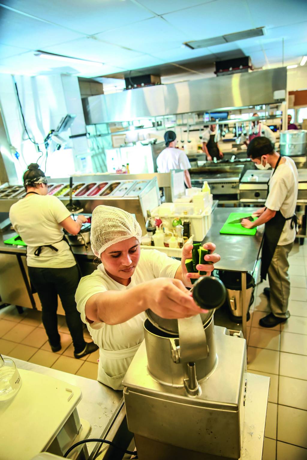 DEVORO_23.JPG Imagem mostra cozinha com cozinheiros de roupas brancas e aventais. Em primeiro plano, uma mulher de touca colocando legumes em aparelho