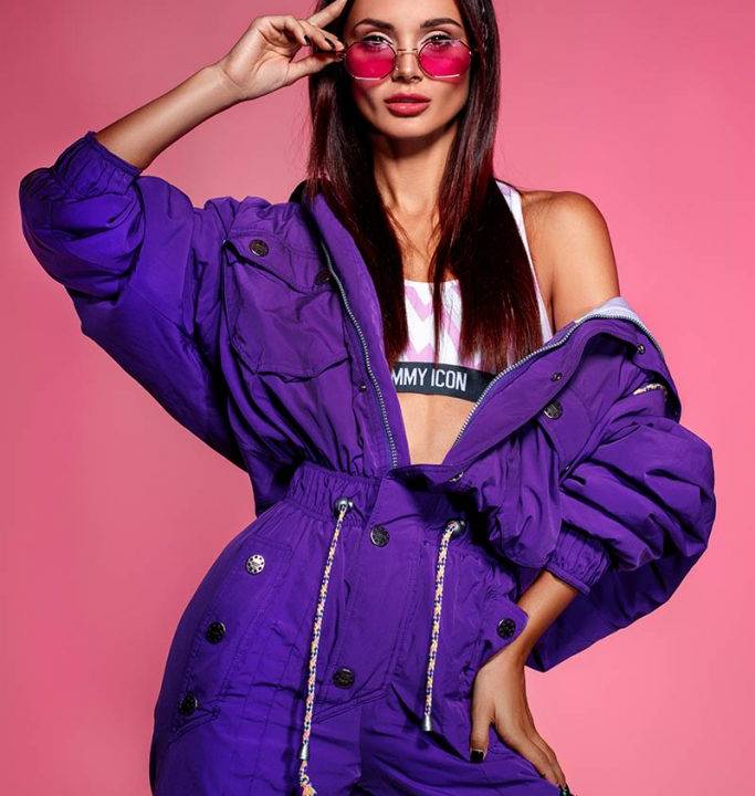 DJ Korolova posa segurando óculos de lentes vermelhas. Ela veste um macacão roxo com cropped branco por baixo. Tem cabelos longos castanhos.