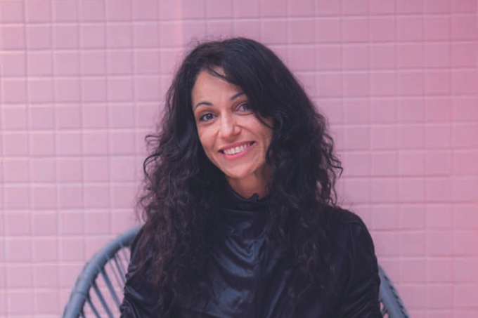 Imagem mostra mulher de cabelo preto e roupa preta sorrindo para a câmera. Ao fundo, uma parede de azulejos rosa.