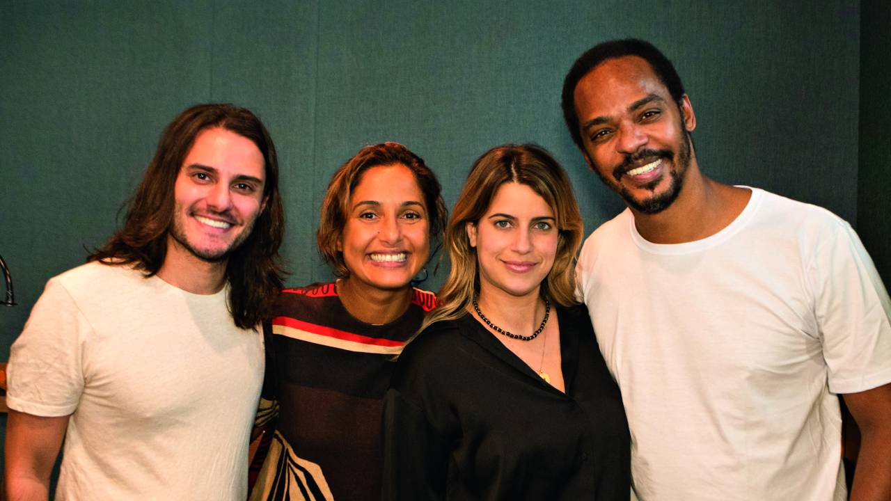 Imagem mostra quatro pessoas abraçadas, sorrindo para a câmera. Ao fundo, uma parede cinza