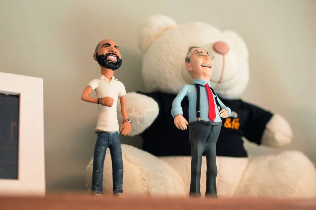 Tomás Covas Miniaturas de Bruno e Mário Covas em cima de uma estante, com um ursinho branco de pelúcia atrás
