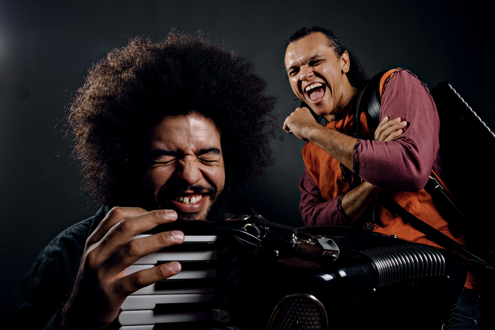 Imagem mostra dois homens rindo. O da esquerda, m primeiro plano, segura uma sanfona