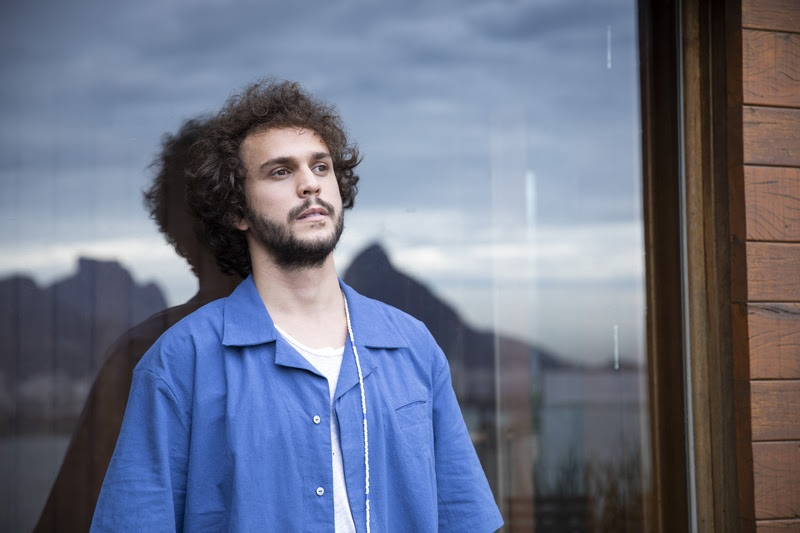 Imagem mostra homem de camisa azul e camiseta branca com as costas apoiadas em vidro. No reflexo, um morro