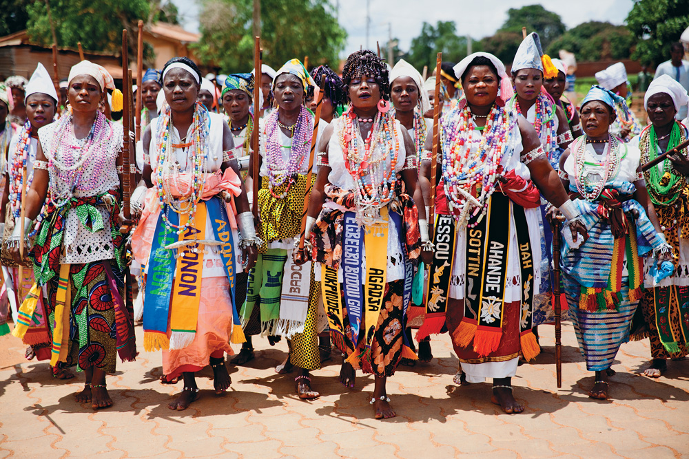 7, Benim, Procissão de sacerdotisas do Dan.jpg Imagem mostra mulheres caminhando em chão de terra, vestindo saias e roupas com diversos adereços coloridos