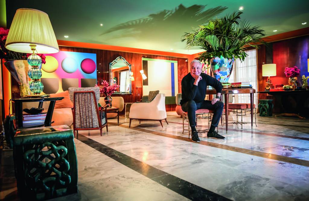 CASACOR_21.JPG Imagem mostra homem sentado em sala colorida, com chão de mármore, poltronas, quadros e um vaso grande ao centro.