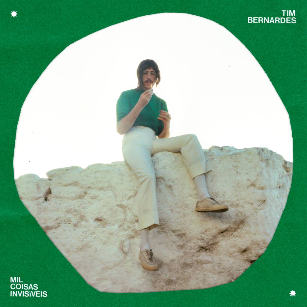 Capa disco tim bernardes Imagem mostra homem de suéter verde e calça branca sentado em mureta de pedra. A imagem é preenchida por um contorno verde.
