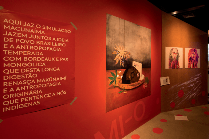 Imagem mostra sala com parede vermelha com uma pintura e um cartaz com um manifesto