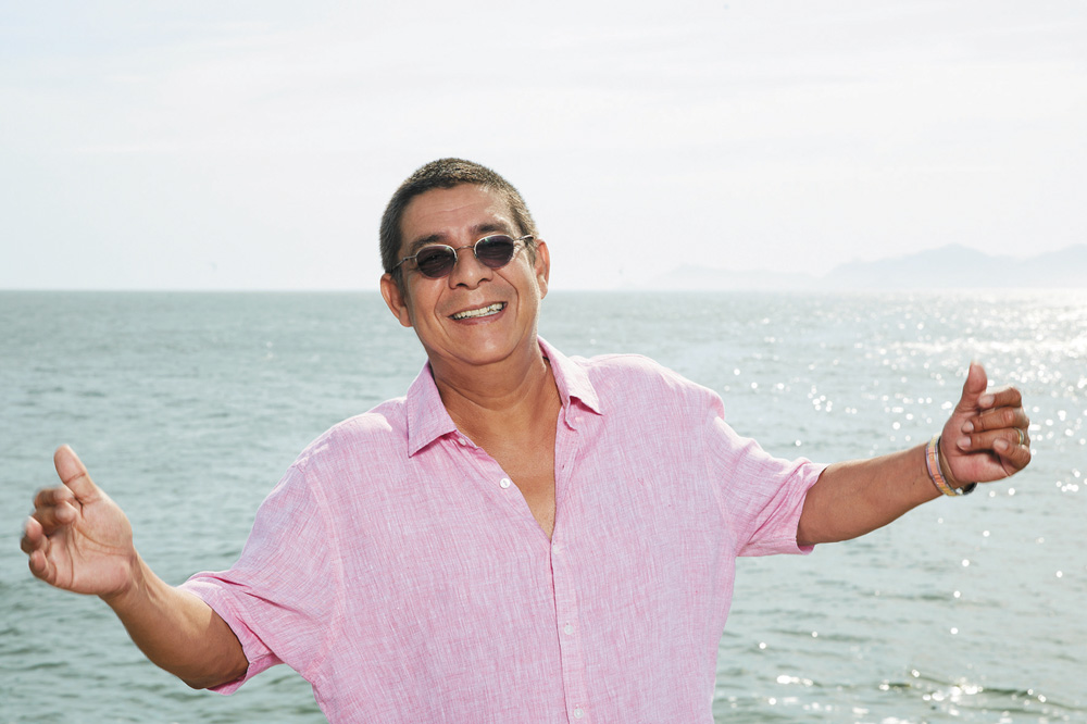 Imagem mostra homem de camisa rosa com os braços abertos, com o mar ao fundo