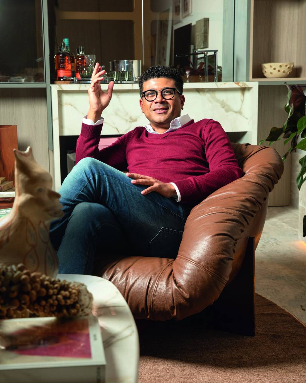 CASACOR 2022 Imagem mostra homem de calça jeans e suéter vermelho sentado em poltrona de couro marrom.