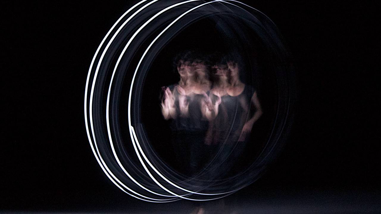 Dançarina parece duplicada no palco. Luzes brancas a circulam em um ambiente escuro