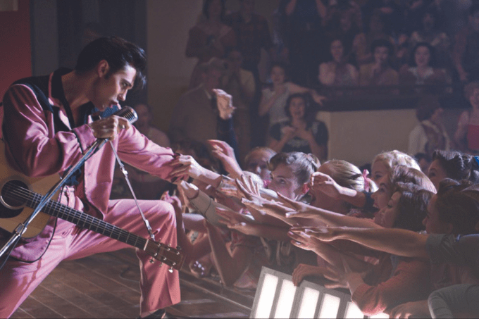 Imagem mostra homem de topete e gel no cabelo em cima de palco, vestindo terno rosa, estendendo a mão para multidão na plateia