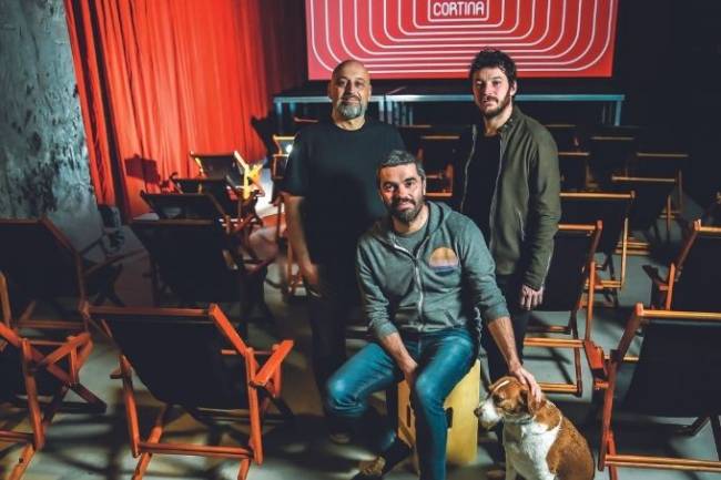 Três homens brancos de barba e cabelos escuros dentro de uma sala de cinema. O que está sentado faz carinho em uma cachorrinha sentada, cujo pelo é uma mescla de branco e caramelo