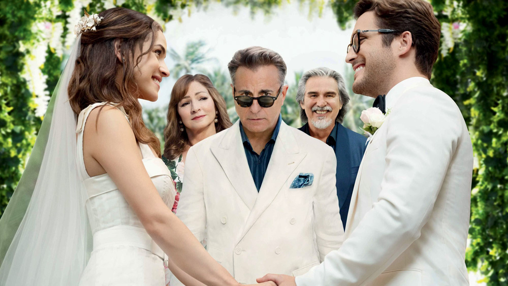 Imagem mostra casal com roupa de casamento com as mãos dadas, frente a frente, e um homem de terno branco ao meio, ao fundo, de óculos escuros, olhando para a câmera