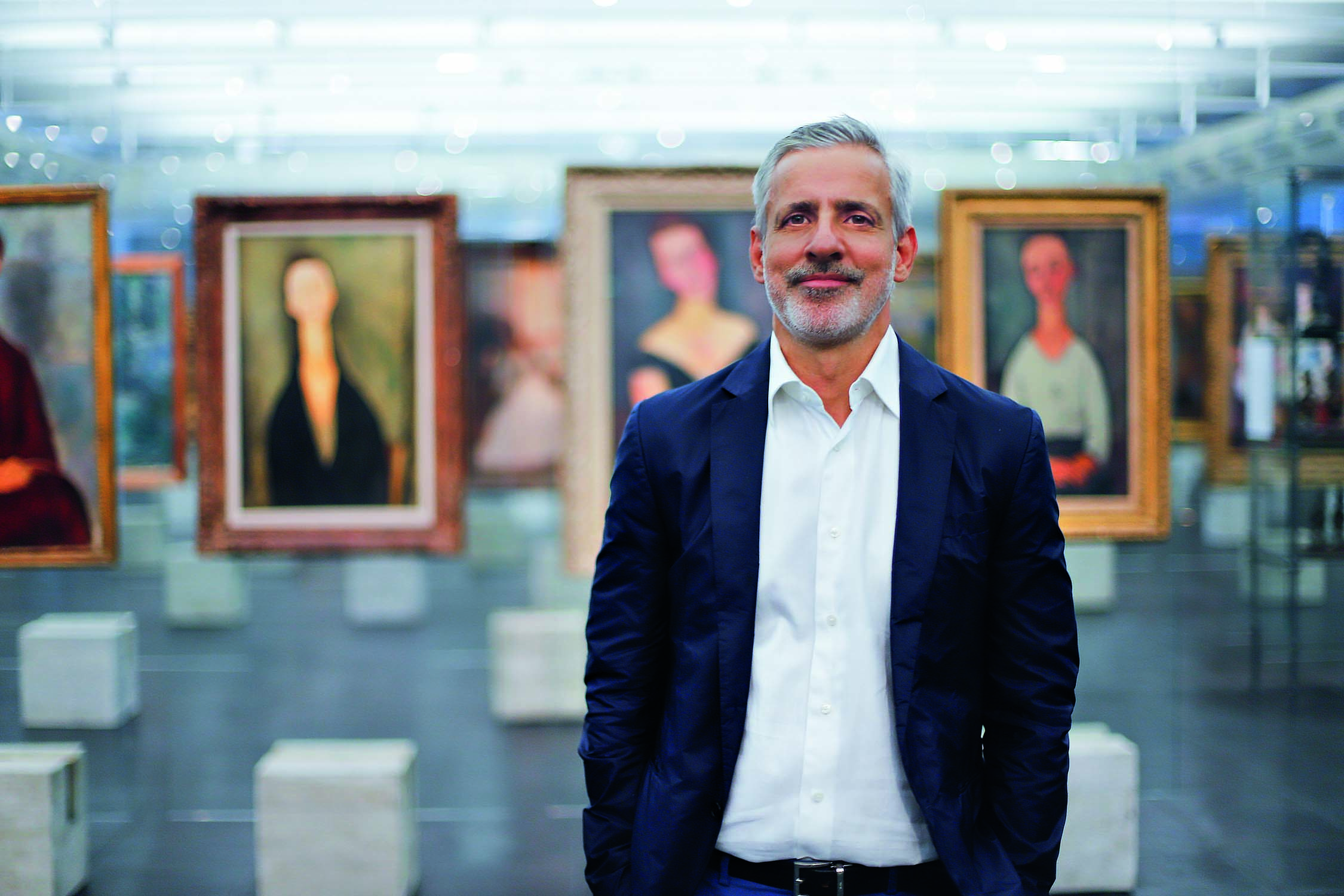 Imagem mostra homem de barba grisalha e camisa e terno em sal&atilde;o de museu. Ao fundo, diversas pinturas.