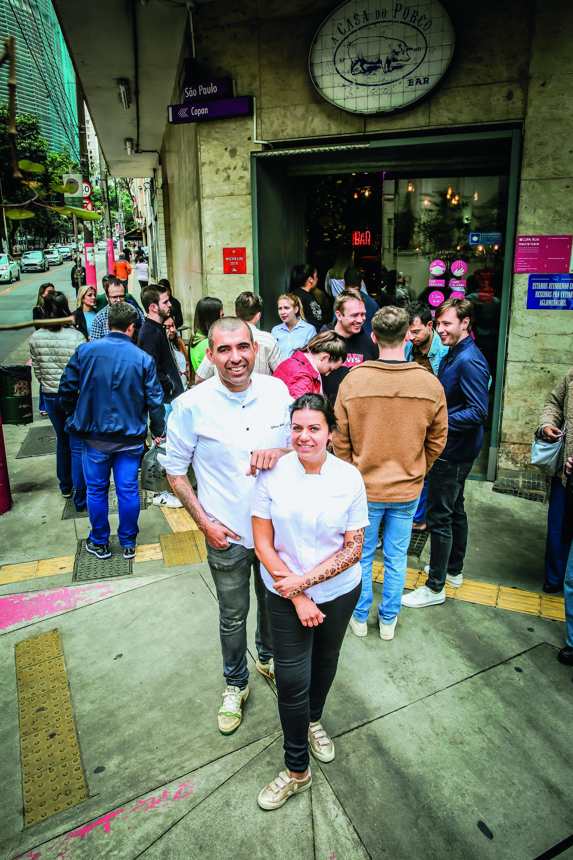 Imagem mostra casal de chefs vestindo roupas brancas em esquina com aglomera&ccedil;&atilde;o na entrada de estabelecimento