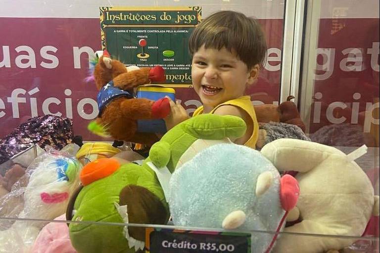 Imagem exibe menino sorridente dentro de máquina repleta de bichinhos de pelúcia coloridos.