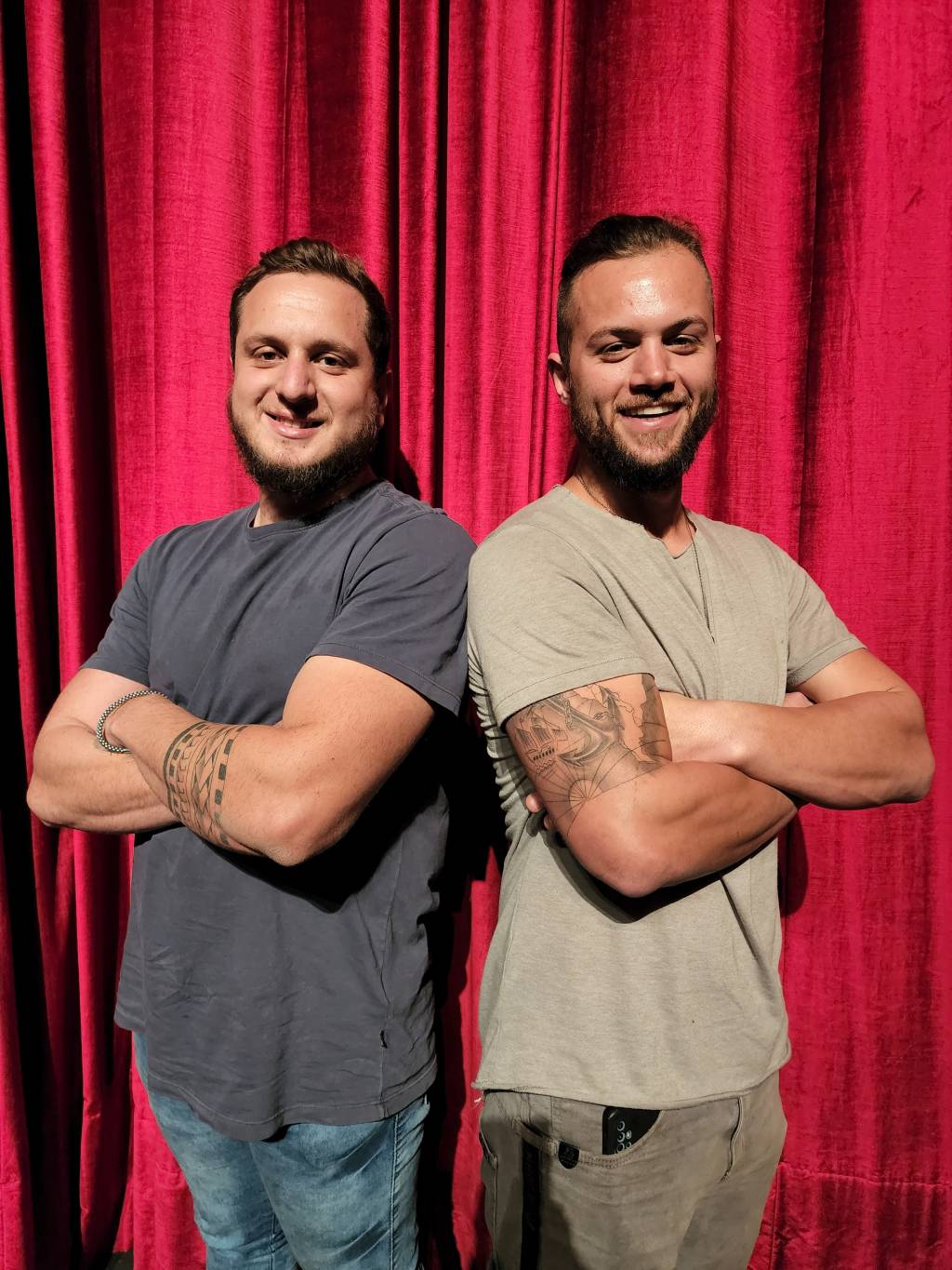 irmãos Lucas e João, Happy Day Circus (MARIANA REDER-DIVULGAÇÃO) (3) Dois homens posam de braços cruzados sorrindo. Ambos vestem camisetas escuras e estão em frente a cortina vermelha.