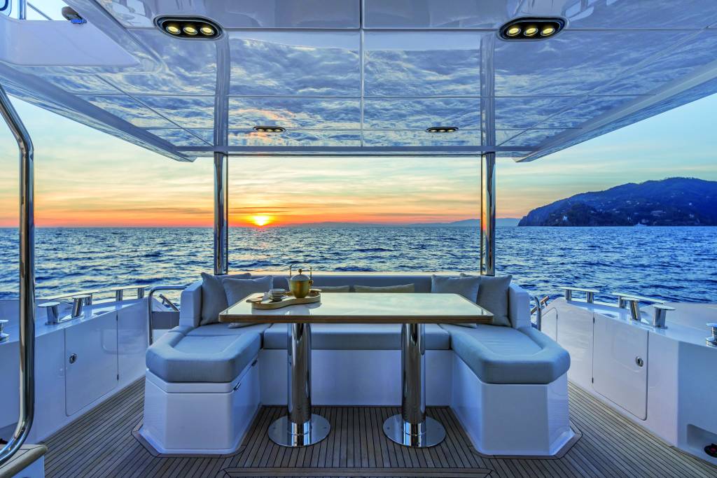 Azimut 62 (Divulgação Azimut Yachts) (3) Foto de mesa com assento azul e branco e mar com horizonte ensolarado ao fundo.