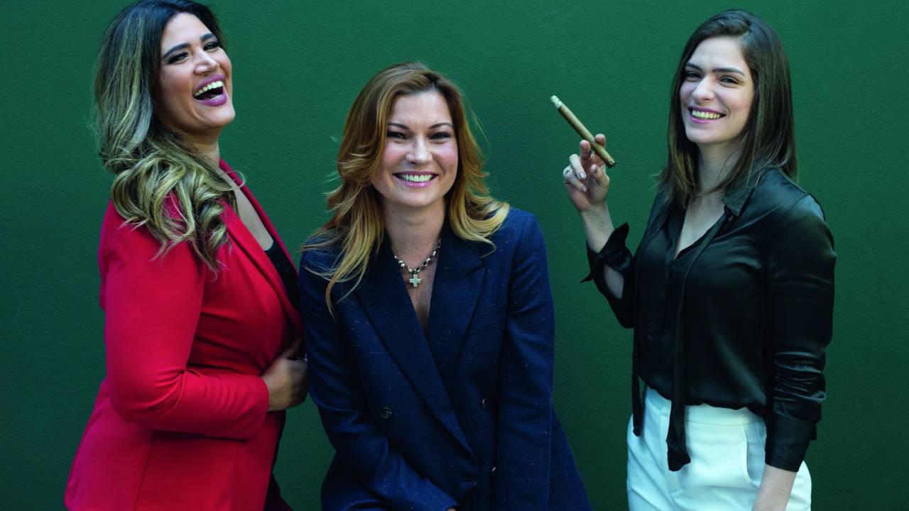 Raquel, Natalie e Cintia posam lado a lado. A partir da esquerda, vestem terninho vermelho, terninho azul escuro e calça branca com blusa preta de seda. A terceira segura um charuto na mão. A do meio aparece sentada e as demais de pé. Todas sorriem. O fundo da parede é todo verde.
