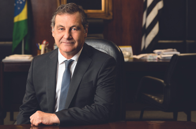 Imagem mostra homem de terno, sorrindo, com uma mão sobre a mesa de madeira