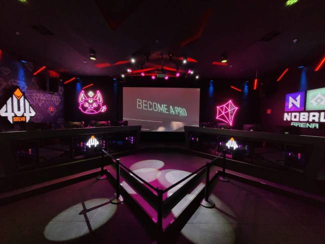 Arena Nobru Imagem mostra palco da Arena Nobru, com duas bancadas com quatro computadores cada, e ao fundo, telão de LED e placas escritas 4UP e Nobru Arena