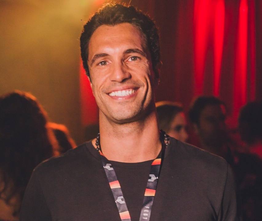 Flavio Sarahyba sorri para a foto e veste camiseta preta com crachá no pescoço.