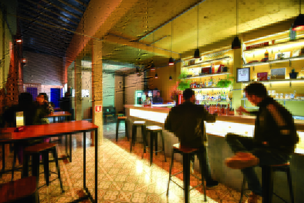 Ambiente interno de bar em que aparece o balcão e duas mesas com banquetas, com dois homens sentados no balcão e duas mulheres próximas às mesas