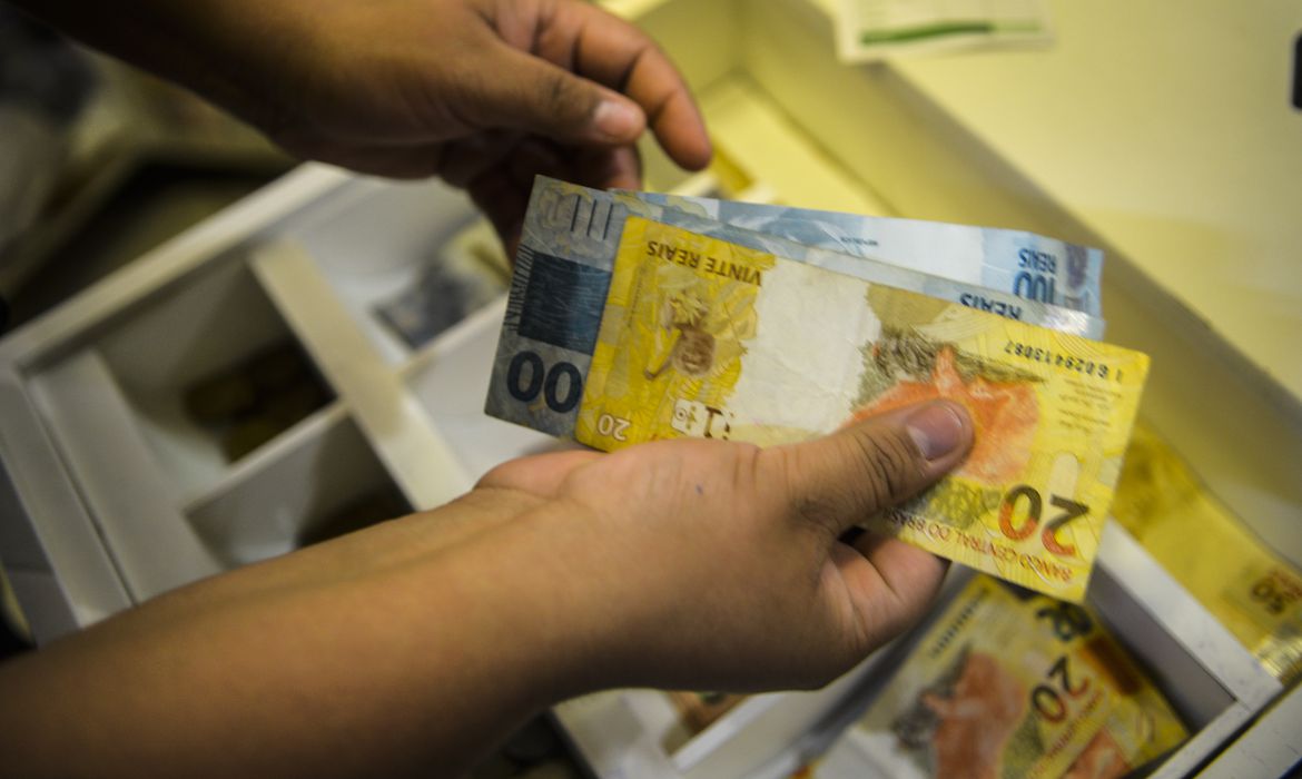 Pessoa segurando uma nota de 20 e outra de 100 diante de caixa registradora