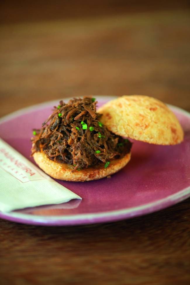 Mesa de madeira com prato cor de rosa, pão de queijo recheado com carne de panela com guardanapo de papel ao lado