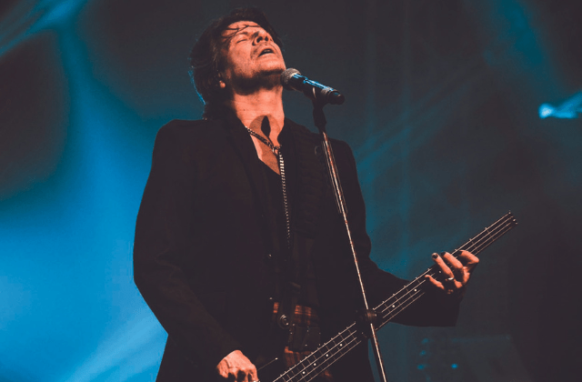 Imagem mostra homem de terno em cima de palco, tocando baixo e cantando em microfone